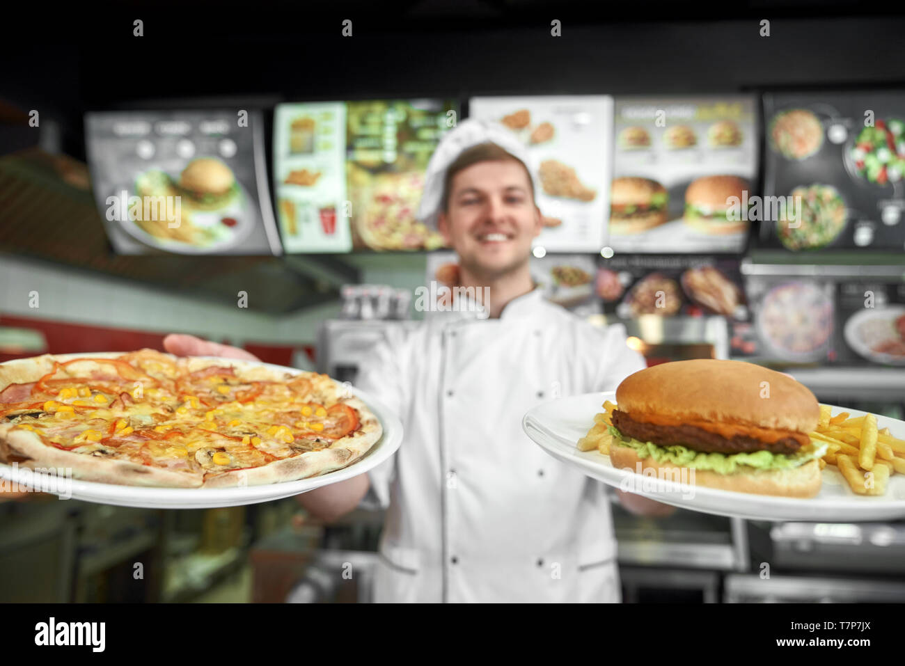 Messa a fuoco selettiva di una gustosa pizza e haumburger con patate libero nelle mani del maschio professionale cucinare in pizzeria. Lo chef felice in bianco uniforme la cottura di fast food nel cafe. Concetto di cibo spazzatura. Foto Stock