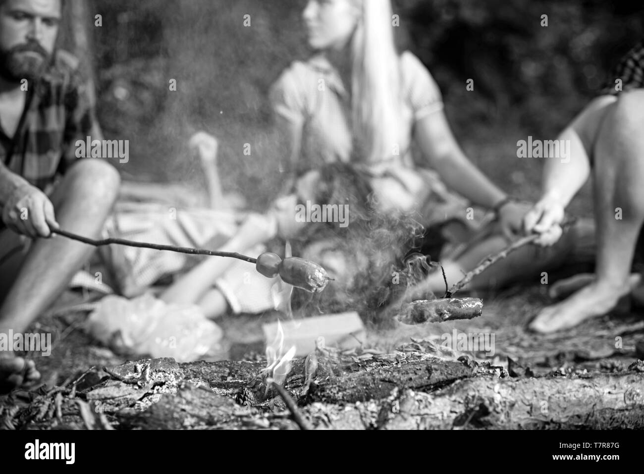 La cottura di salsicce su bastoni su fiamme di fuoco, la notte in campeggio in estate. Gruppo di amici seduti davanti al falò Foto Stock