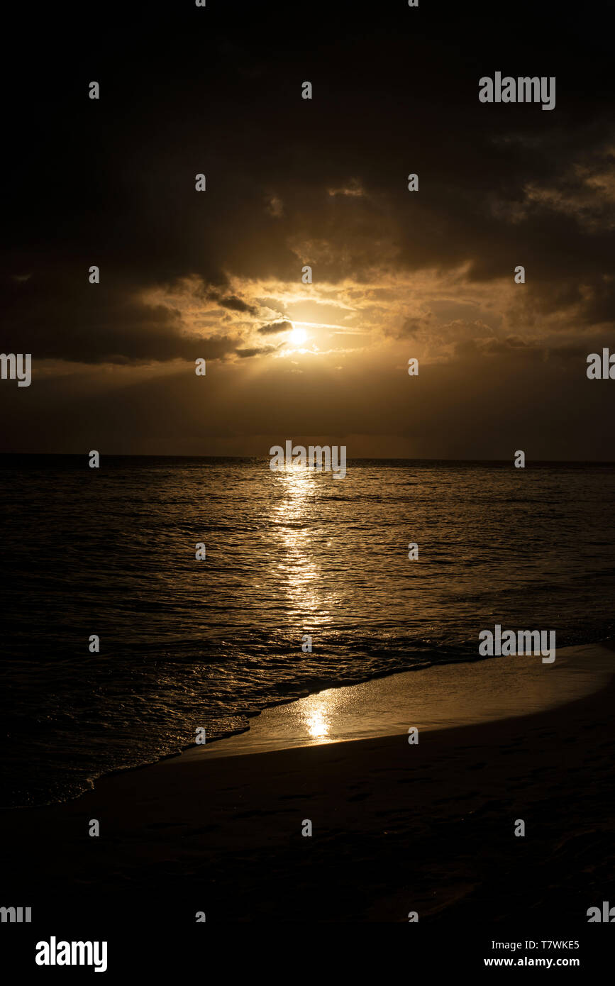 Sole che tramonta dietro le nuvole sopra la riva del mare wioth onde che lambiscono contro una spiaggia sabbiosa Foto Stock