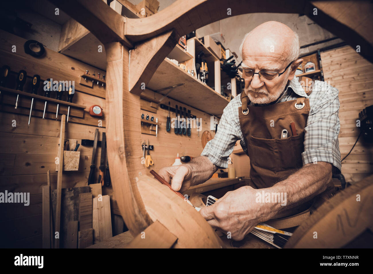 Un vecchio falegname prende le misure ad una sedia per essere ripristinato, un workshop con gli strumenti in background. Foto Stock