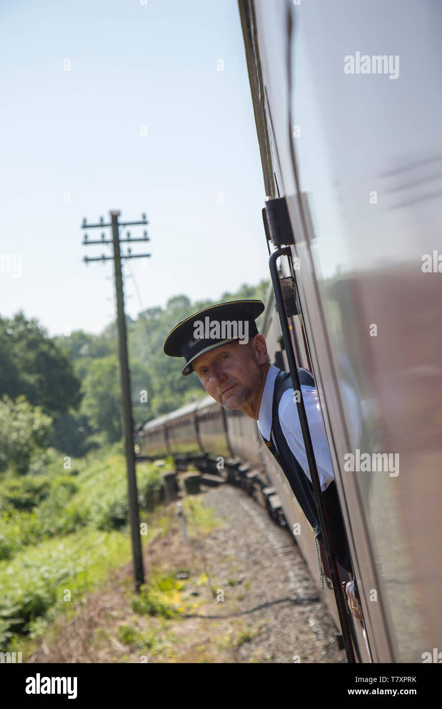 Close up ritratto di elegantemente vestiti di protezione del treno in berretto, a bordo vintage UK treno a vapore, proteso al di fuori della carrozza ferroviaria finestra guardando fuori. Foto Stock