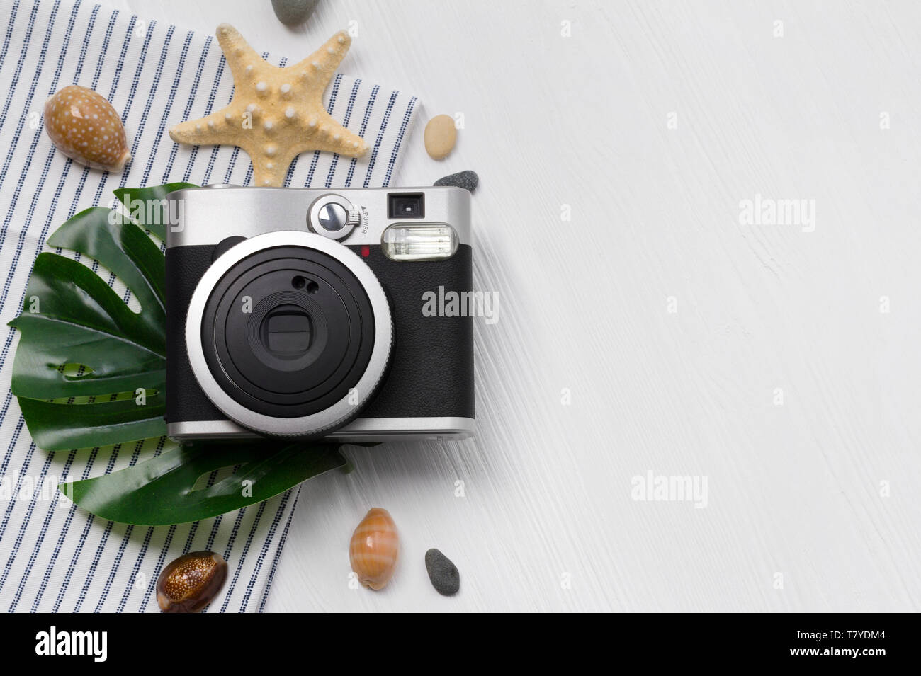 Fotocamera con estate assesories: leav monstera, stella pesce e asciugamano, conchiglie e pietre di colore bianco su un tavolo di legno. Concetto di viaggio. Foto Stock
