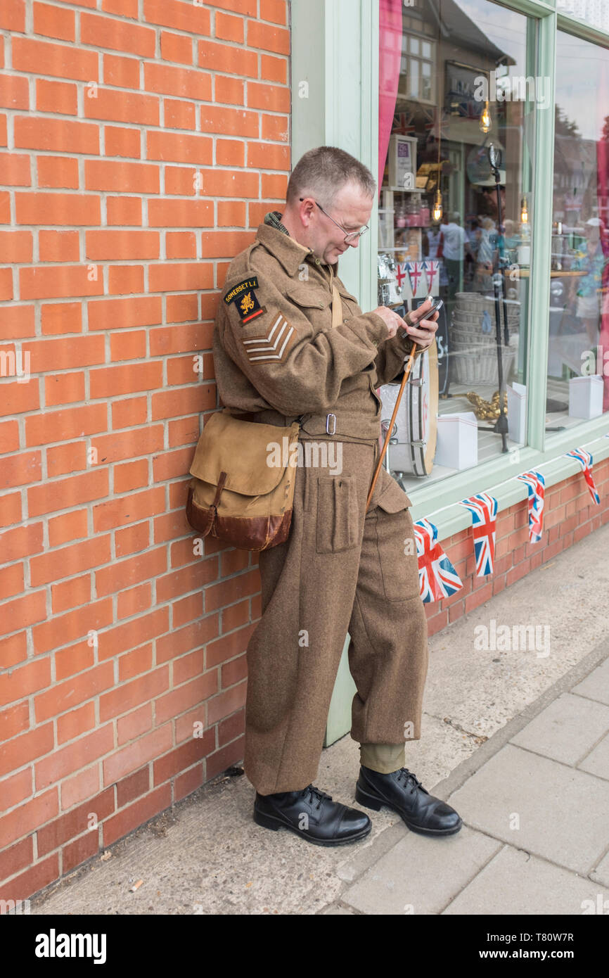 Woodhall Spa 1940s guerra mondiale due festival rievocazione weekend, soldato in WW2 battaglia vestito appoggiato contro una parete utilizzando un telefono intelligente. Foto Stock