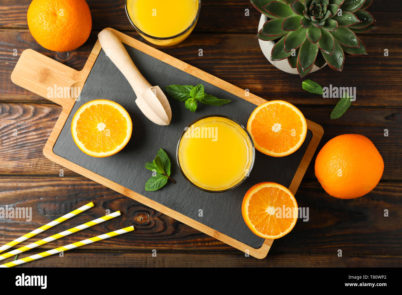 Tagliere, succo di arancia, centrifuga in legno, menta, arancia, tubicino e piante succulente sul tavolo di legno, vista dall'alto. Fresca bevanda naturale e frutta Foto Stock