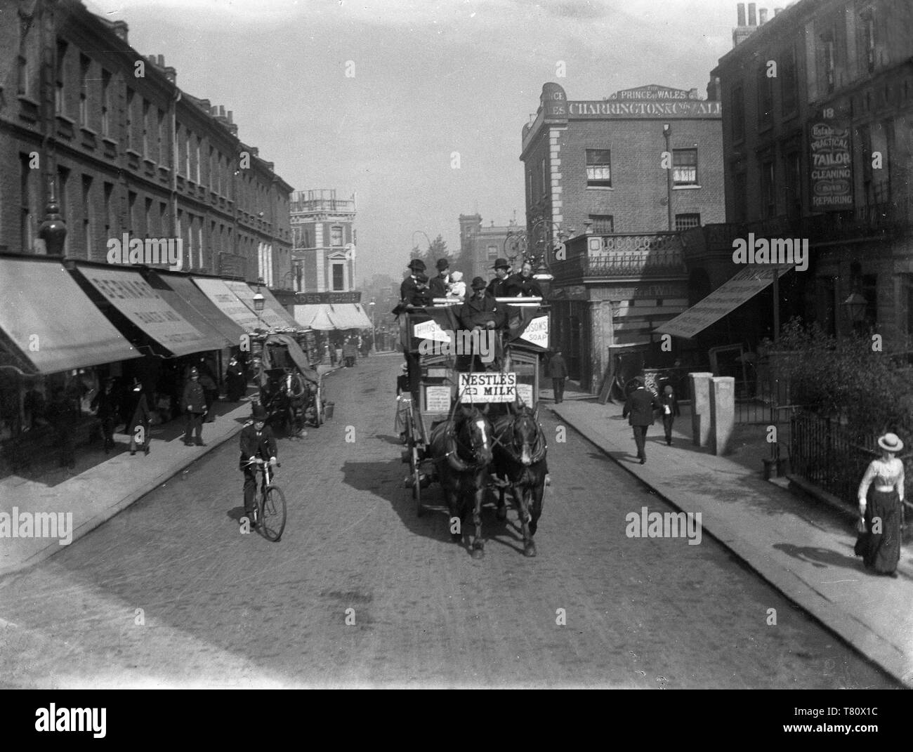 A Londra nel periodo Vittoriano scena di strada di un cavallo e bus e altre persone di andare circa le loro attività quotidiane, inclusi un poliziotto, uomo sulla moto e elegantemente vestito donna, con una discesa di negozi a baldacchino e il Principe di Galles Public House. Nota l'annuncio per Nestle il latte sul bus. Foto di Tony Henshaw Foto Stock