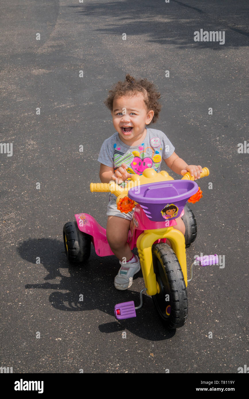 Vadnais Heights, Minnesota. Un anno e mezzo di lavoro vecchi bi-razziale ragazza cavalcare i suoi veicoli a tre ruote con una faccia felice. Foto Stock