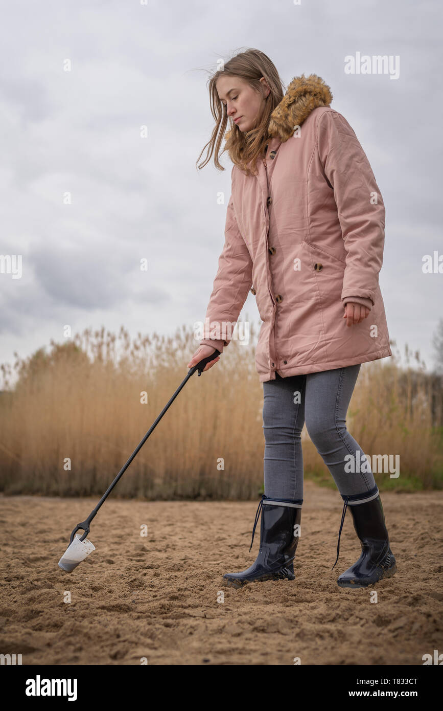 Donna in stivali di gomma raccoglie i rifiuti in plastica su una spiaggia con una pinza Foto Stock