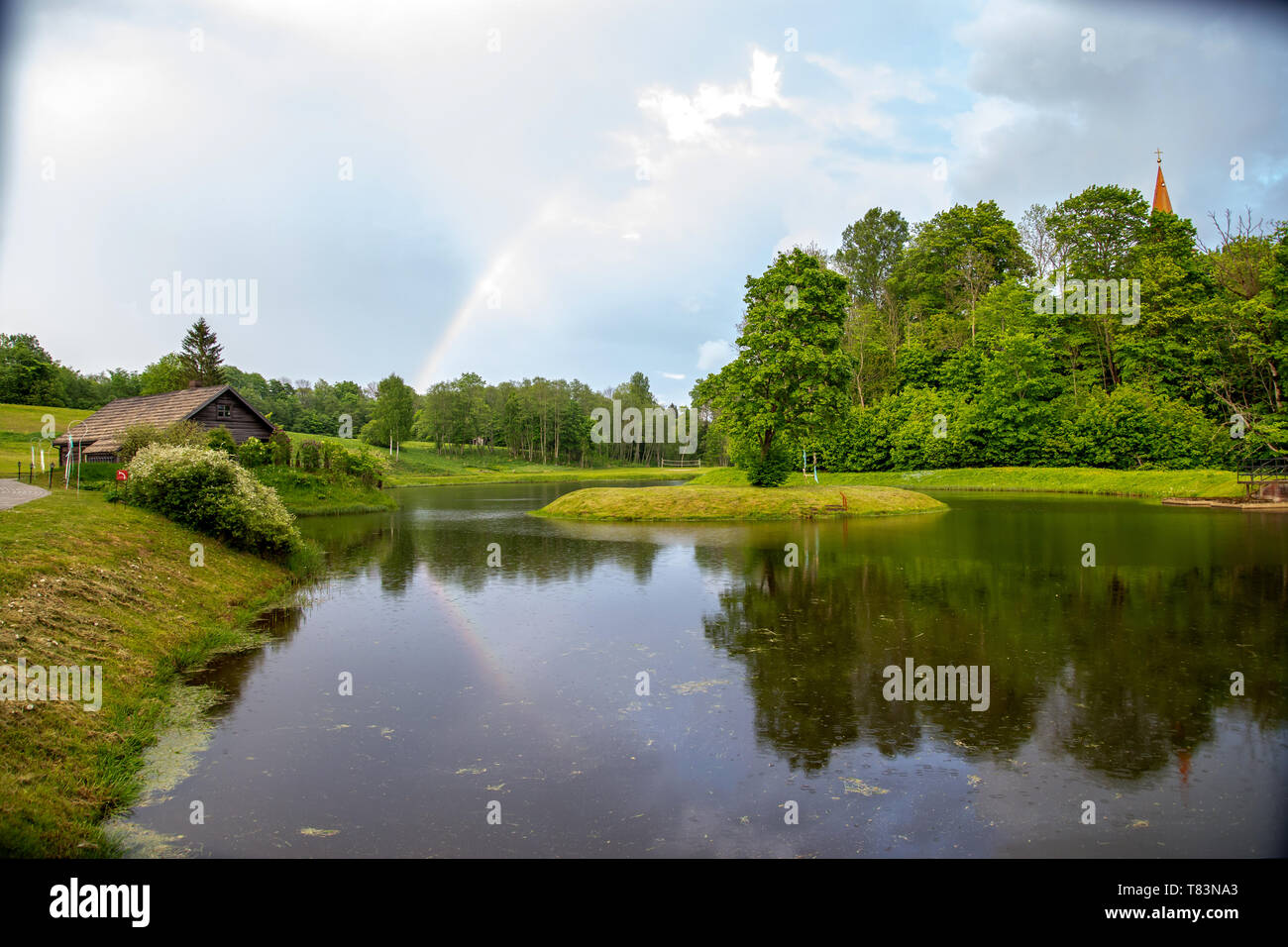 Rainbow oltre lo stagno, alberi e casa in legno sul giorno di estate. Rainbow e alberi riflesso nell'acqua, Lettonia. Foto Stock