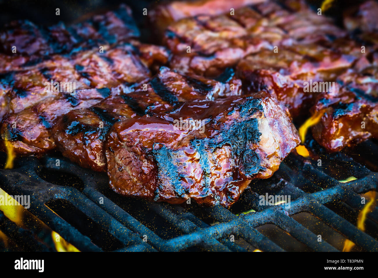 Carni bovine disossate nervature grigliare su fiamme con aggiunta di salsa barbecue. Estrema profondità di campo con sfondo sfocato con focus sulla parte anteriore della carne. Foto Stock