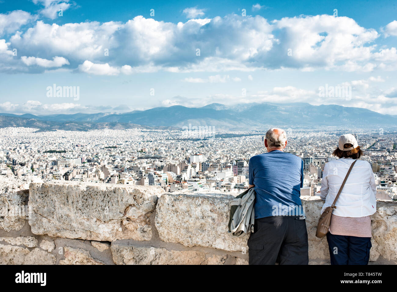 Sito del Patrimonio Mondiale UNESCO dell'Acropoli Paesaggio urbano di Atene Turisti Grecia | ATENE — Due visitatori si appoggiano a un'antica muraglia di pietra sull'Acropoli, godendo di una vista panoramica su Atene che si estende verso le montagne che circondano il bacino dell'Attica. La roccia ha servito come punto di osservazione sulla città per più di 2.500 anni, e la sua altitudine di circa 512 piedi (156 m) sopra il bacino circostante offre uno dei panorami più completi della moderna capitale greca. Foto Stock