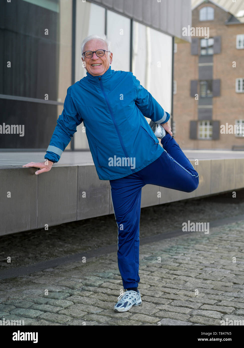 Senior uomo in tuta facendo esercizi di riscaldamento sulla strada di ciottoli, ritratto, Copenaghen, Hovedstaden, Danimarca Foto Stock