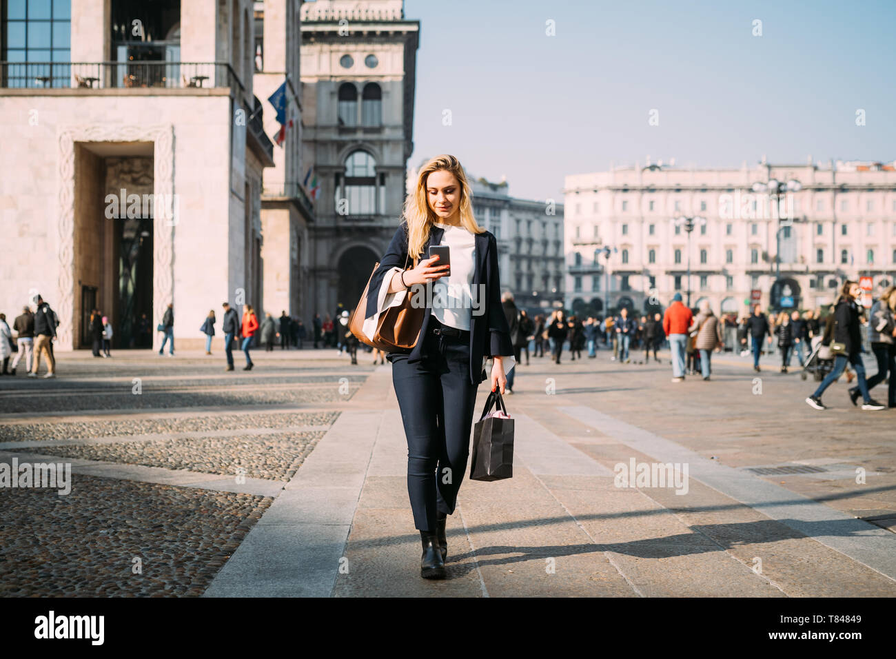 Giovane turista femminile con borse per lo shopping passeggiando e guardando lo smartphone nel city square, Milano, Italia Foto Stock