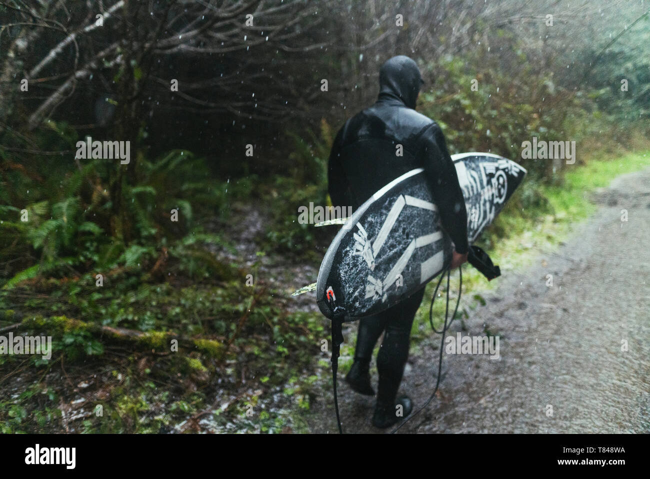 Giovane maschio surfer in wetsuit a piedi lungo puddled sporco della pista sotto la pioggia, in vista posteriore, Arcata, California, Stati Uniti Foto Stock
