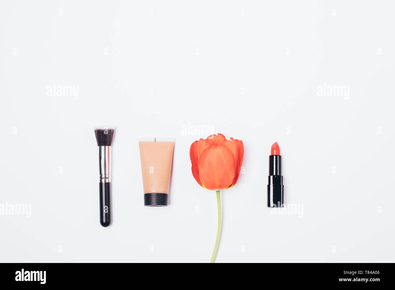 Prodotti cosmetici per classic basic make-up, piatta concetto. Vista superiore spazzola, crema fondotinta e rossetto rosso accanto al tulip flower su sfondo bianco. Foto Stock