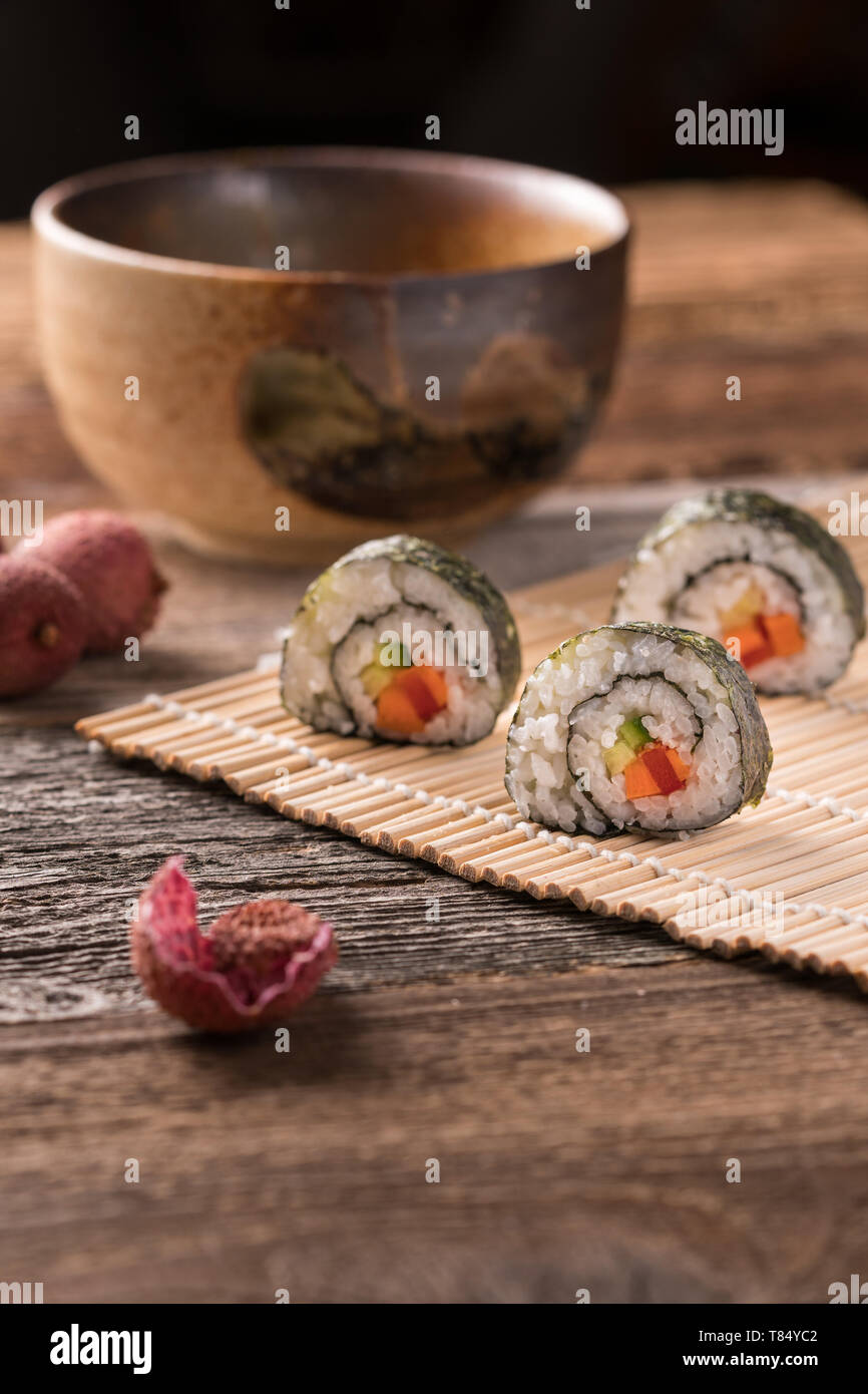 Tre maki sushi rotoli sul tappetino di bambù sul tavolo di legno, tradizionali asiatici tee ciotola, litchi e litchi buccia - angolo di visualizzazione, sfondo sfocato, verticale Foto Stock