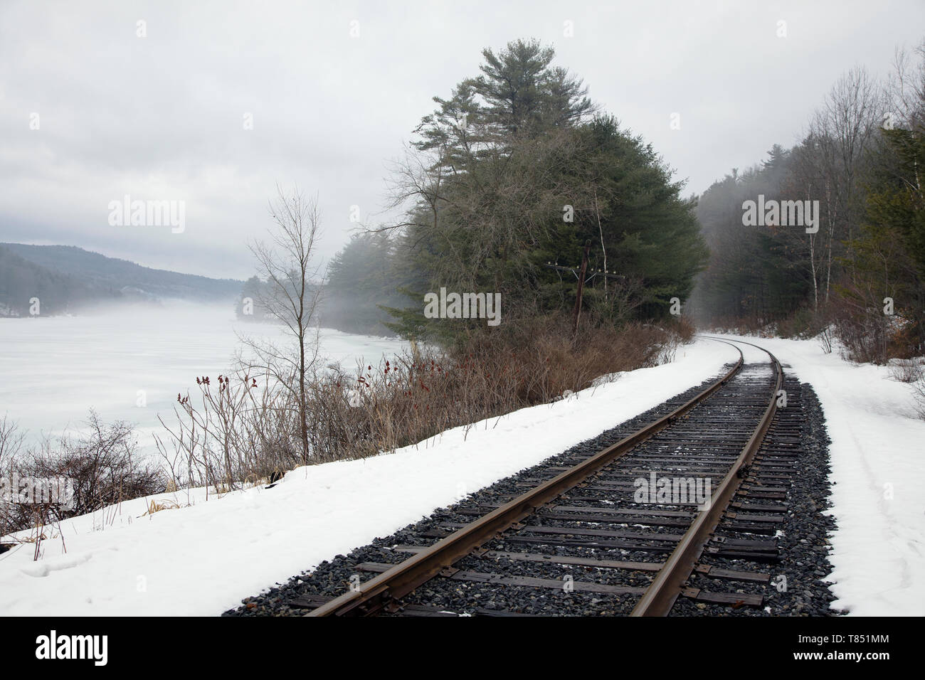 Un binario ferroviario, treno tracce nella neve, da un lago nel paesaggio del Vermont, di nebbia che salgono dal acqua, montagne in distanza. Foto Stock