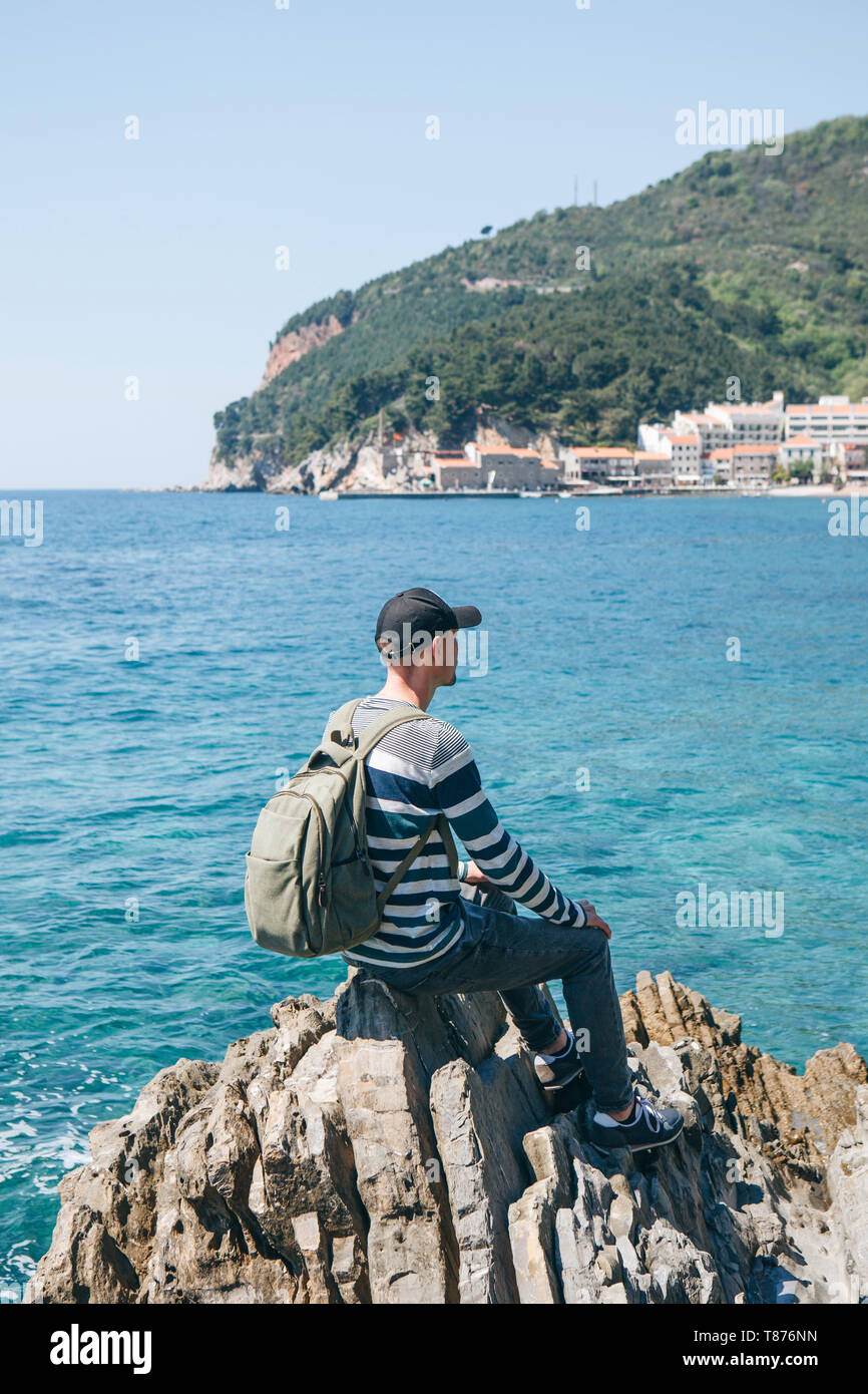 Un turista con lo zaino da solo si trova su di una scogliera sul mare e guarda nella distanza alla vista di Petrovac in Montenegro. Foto Stock