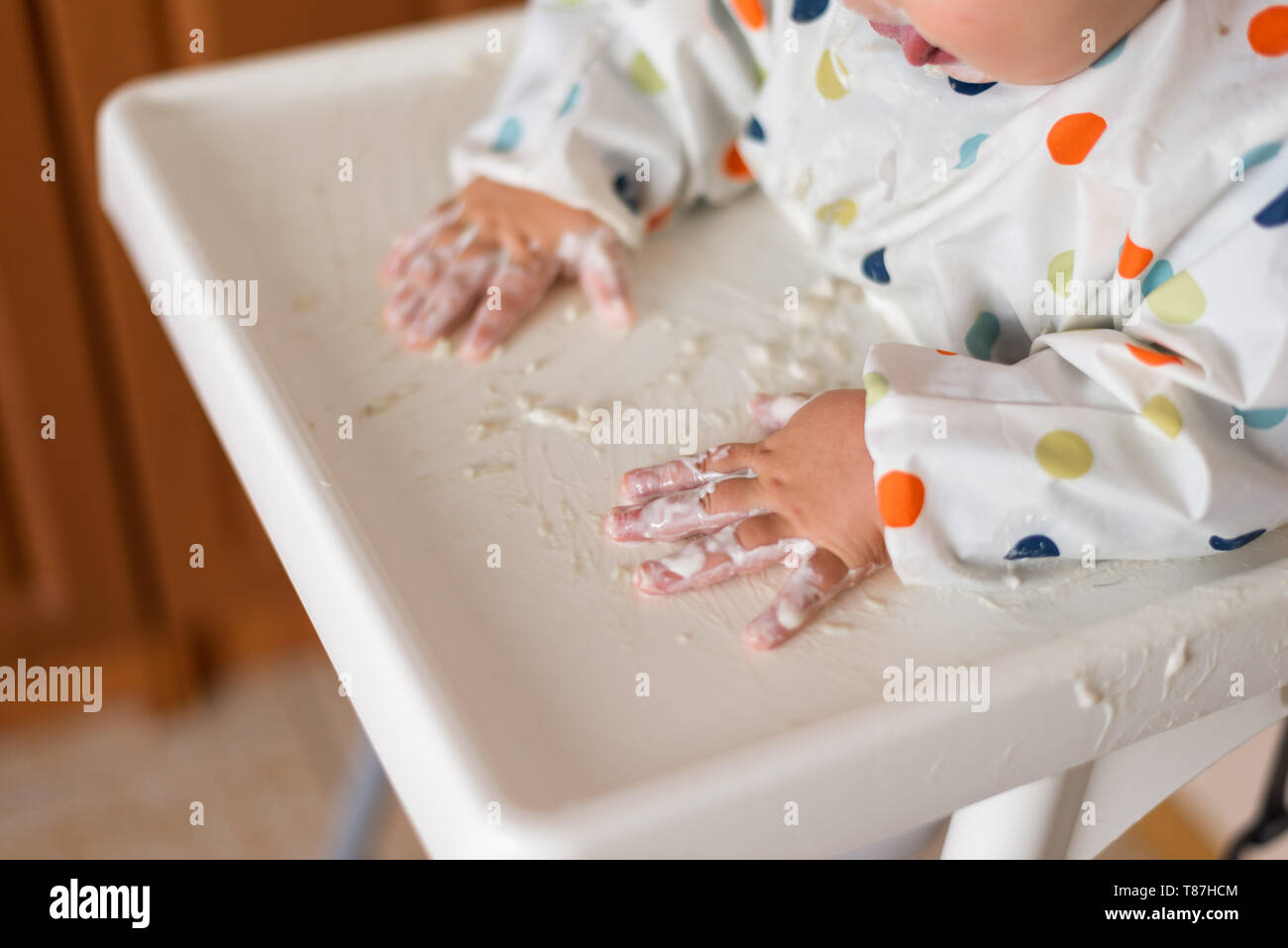 Una bambina in una t-shirt e la piastra seduto in una sedia per il bambino a mangiare con le mani i cereali con aggiunta di yogurt Foto Stock