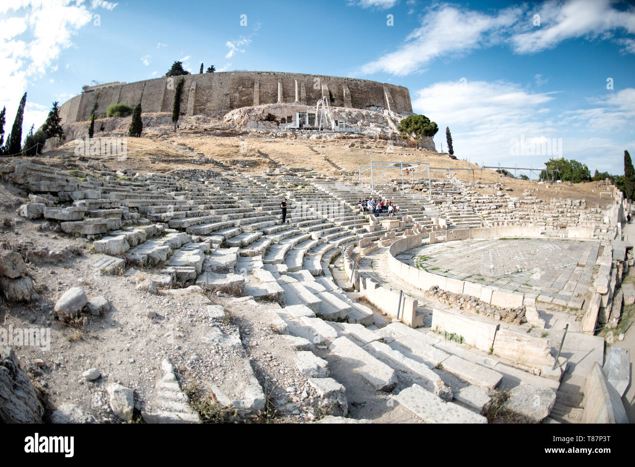 Odeion di Erodiano Attico Antico teatro romano Collina dell'Acropoli Atene Grecia | ATENE — I visitatori siedono tra i gradini di pietra curvi dell'Odeion di Erodiano Attico, il teatro dell'epoca romana costruito nel versante sud-occidentale dell'Acropoli. Il ricco senatore Erodiano Attico costruì il luogo nel 161 d.C. in memoria di sua moglie, e i suoi posti a sedere in marmo restaurati ospitano ancora spettacoli durante il Festival annuale di Atene, sotto la cima fortificata coronata dal Partenone. Foto Stock