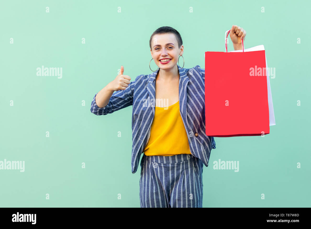 Grande negozio. Ritratto di successo della bellissima con capelli corti giovane donna in tuta a strisce in piedi, azienda red shopping bag e mostrando pollice in alto. Ind Foto Stock