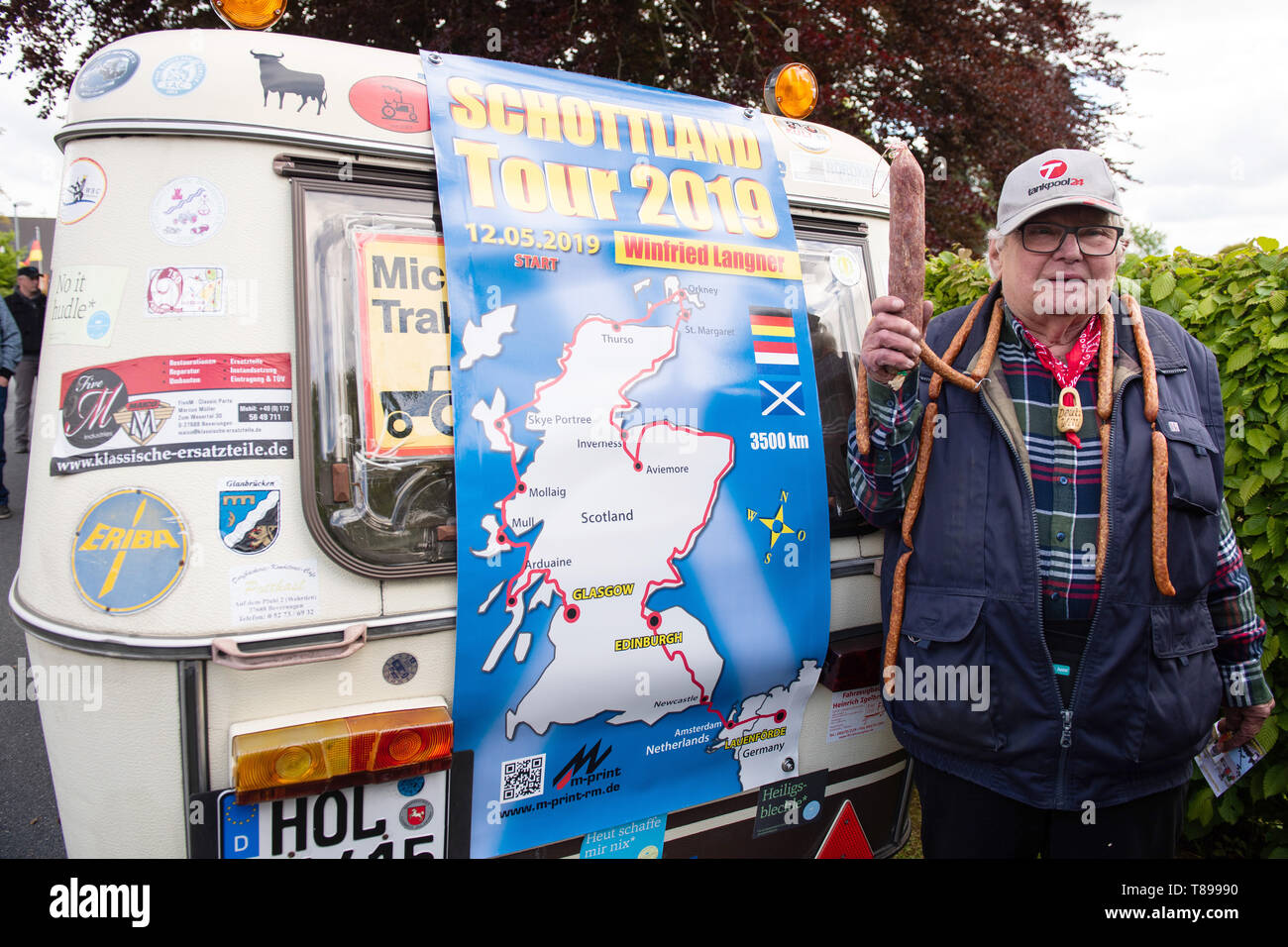 Lauenförde, Germania. Il 12 maggio 2019. Il titolare di pensione o di rendita Winfried Langner, noto come 'Deutz-Willi', si trova prima della partenza con un Mettwurst nella sua mano sul suo rimorchio. 'Deutz-Willi' inizia con il suo trattore Deutz costruito nel 1961 e la sua piccola carovana in Scozia. Egli sarà probabilmente sulla strada per cinque mesi. Foto: Swen Pförtner/dpa Credito: dpa picture alliance/Alamy Live News Foto Stock