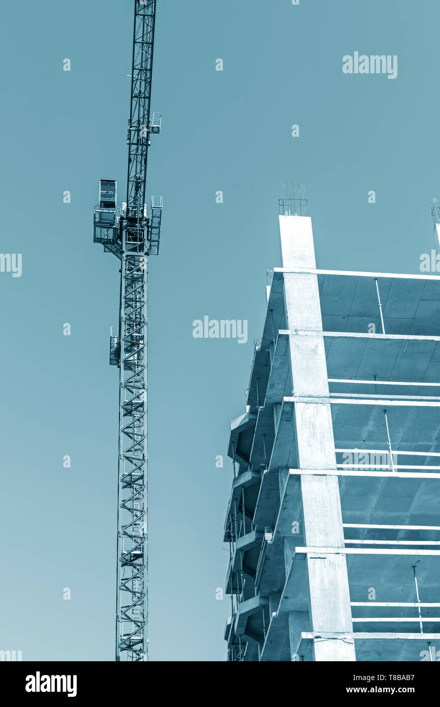 Alta gru a torre in piedi vicino a blocco città edificio in costruzione contro il cielo blu sullo sfondo Foto Stock