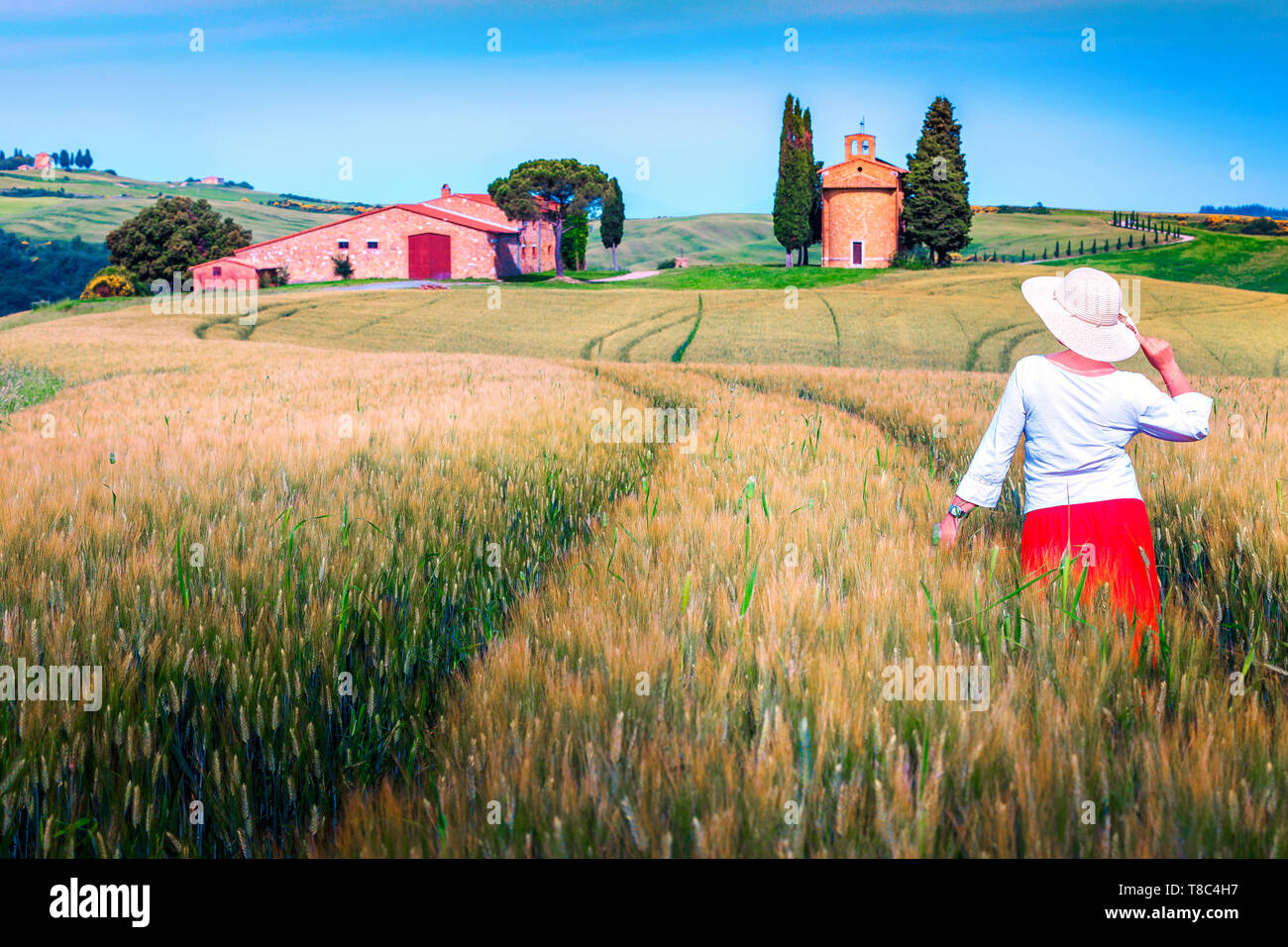 Bella donna felice in rosso mantello e cappello di paglia godendo della libertà. Paesaggio rurale con campi di grano e cappella di Vitaleta, Toscana, Italia, Europa Foto Stock