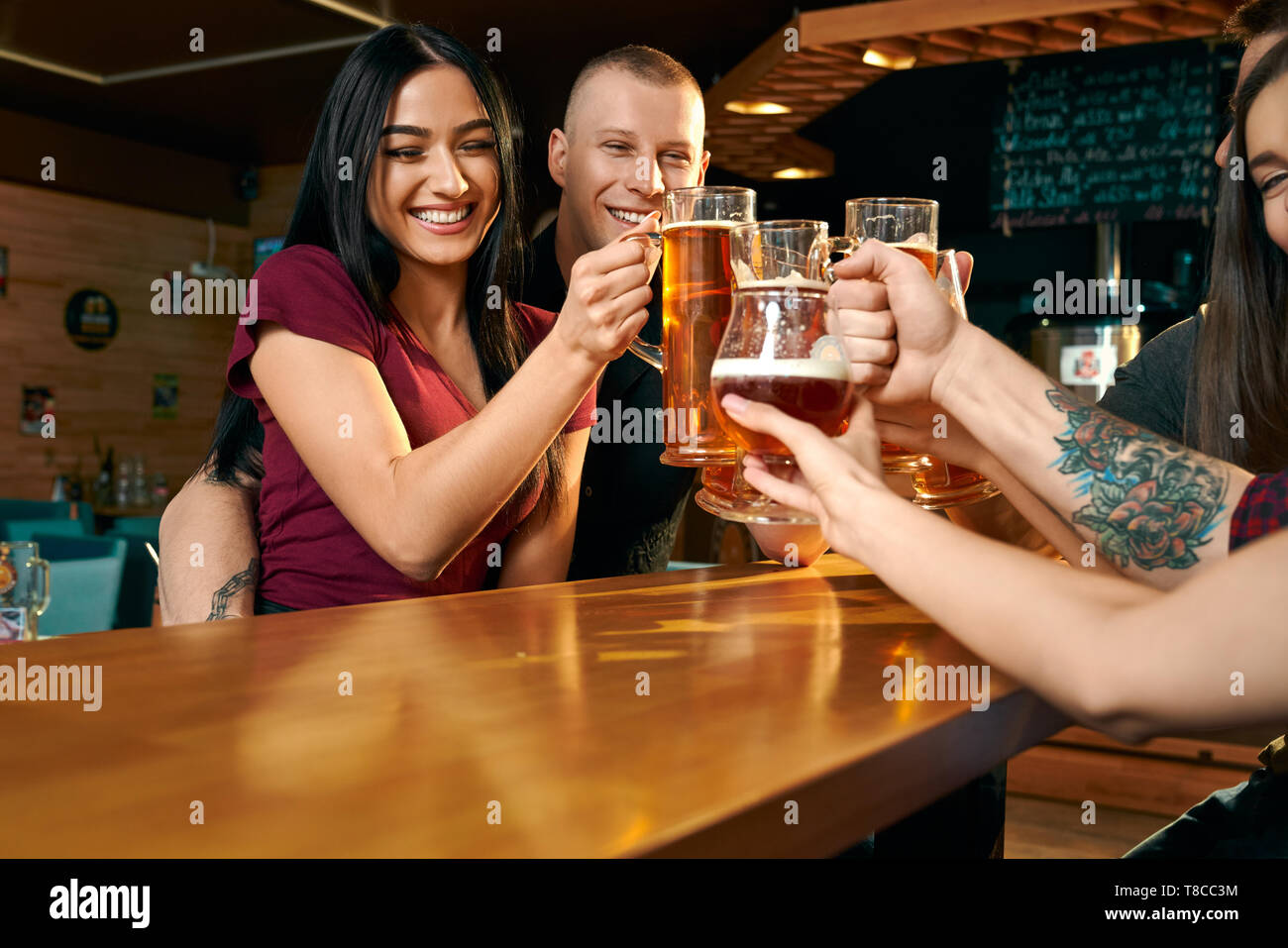 Vista dal lato del felice l'uomo e la donna che abbraccia ogni altra e tostatura con gli amici nel pub. Azienda bere birra, ridendo e godere del tempo libero insieme. Concetto di felicità e di bevanda. Foto Stock