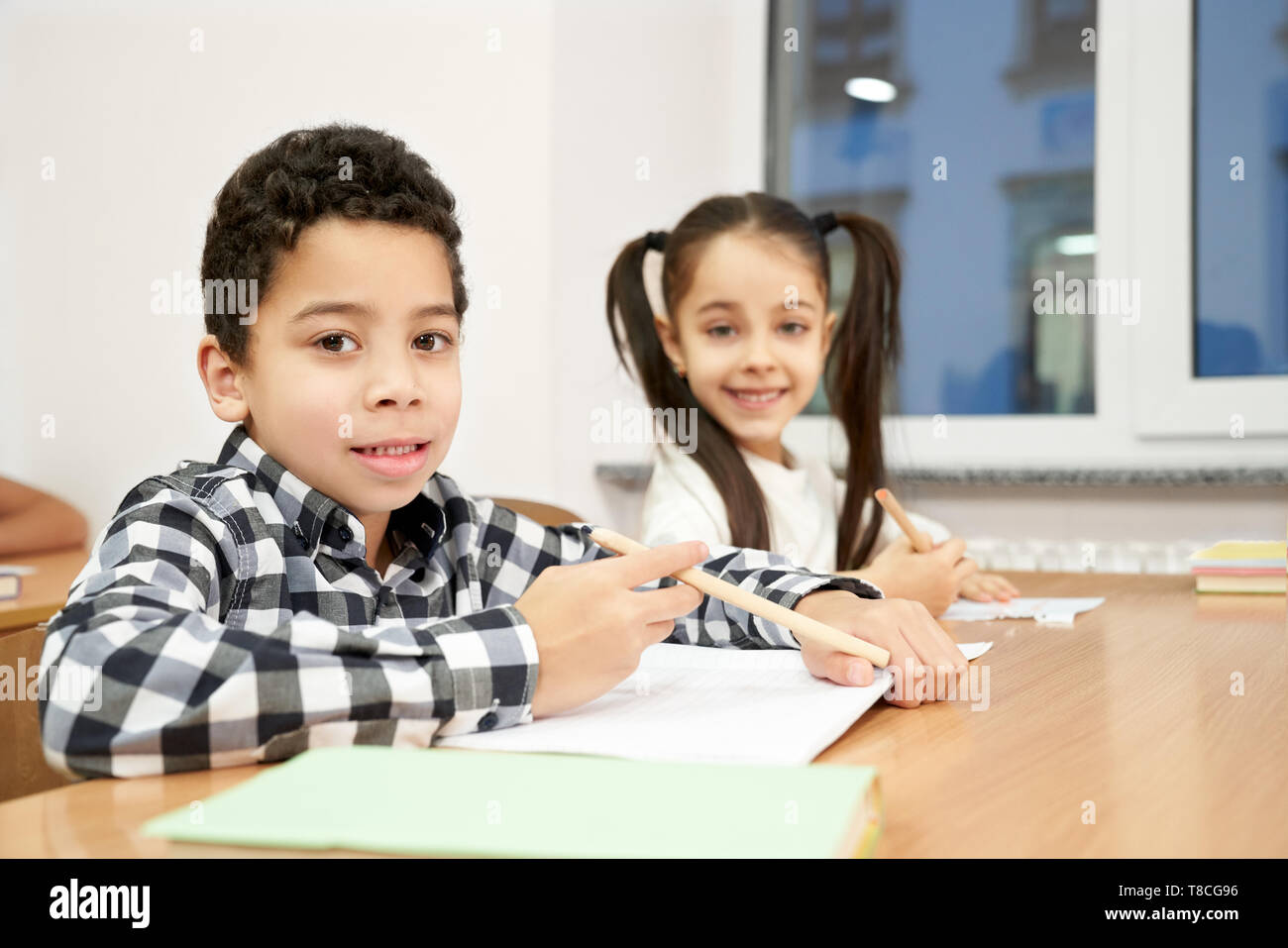Allievo della Scuola primaria seduto alla scrivania, tenendo matite, guardando la telecamera, in posa. Allegro un ragazzo e una ragazza, scolari apprendimento, scrittura in copybook in aula. Foto Stock