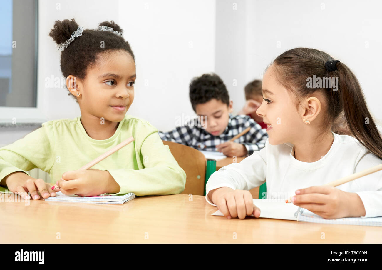Carino, piuttosto studentesse seduto alla scrivania in aula, tenendo in mano la penna il, guardando ogni altra posa. Gli alunni della scuola primaria in scrittura copybook sulla lezione. Foto Stock