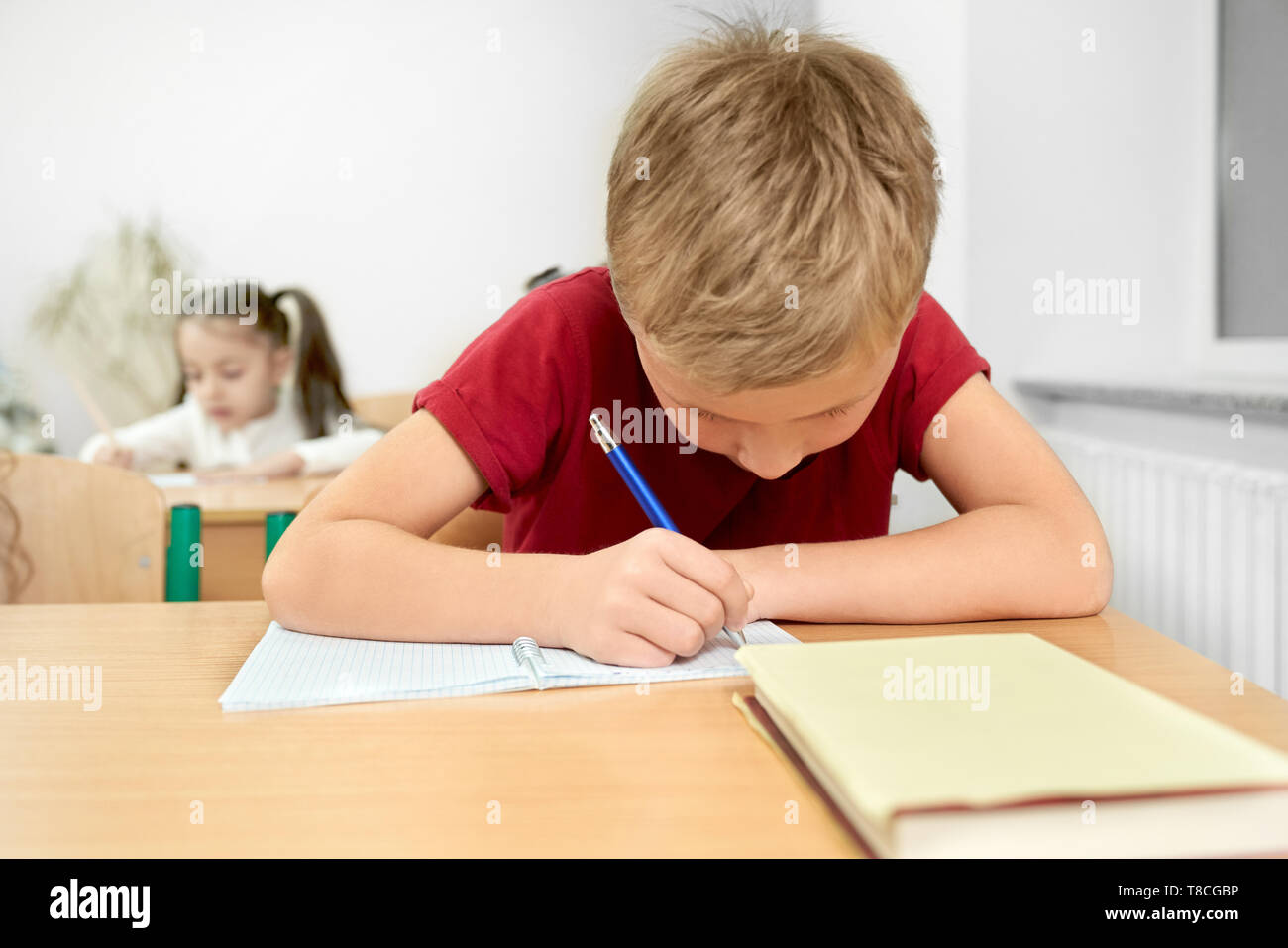 Giovani schoolboy in rosso di t-shirt guardando giù, seduti alla scrivania e scrivere con la penna in copybook. Pupilla concentrato, adolescente di scuola primaria a tavola in aula. Foto Stock