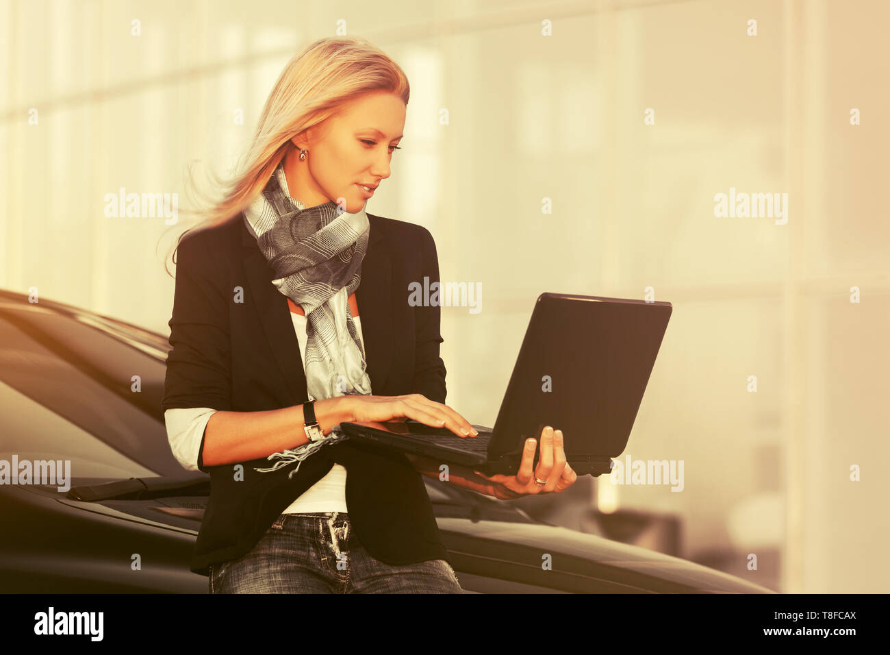 Moda giovane donna di affari con computer portatile seduta sul suo elegante auto modello femminile indossando abito nero giacca grigio e sciarpa per esterno Foto Stock