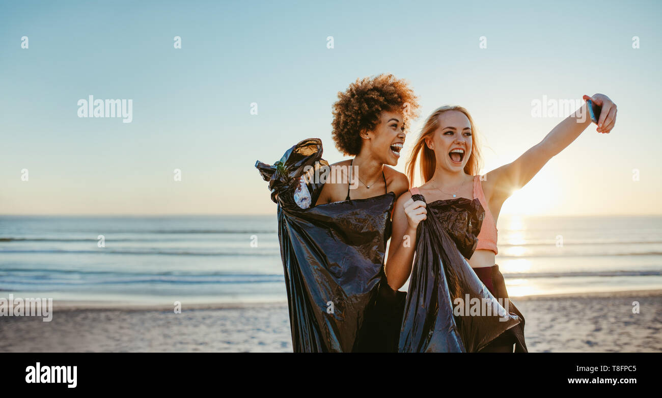 Volontari di sesso femminile con sacchetto di immondizia tenendo selfie sulla spiaggia dopo aver fatto il cestino tag challenge. Foto Stock