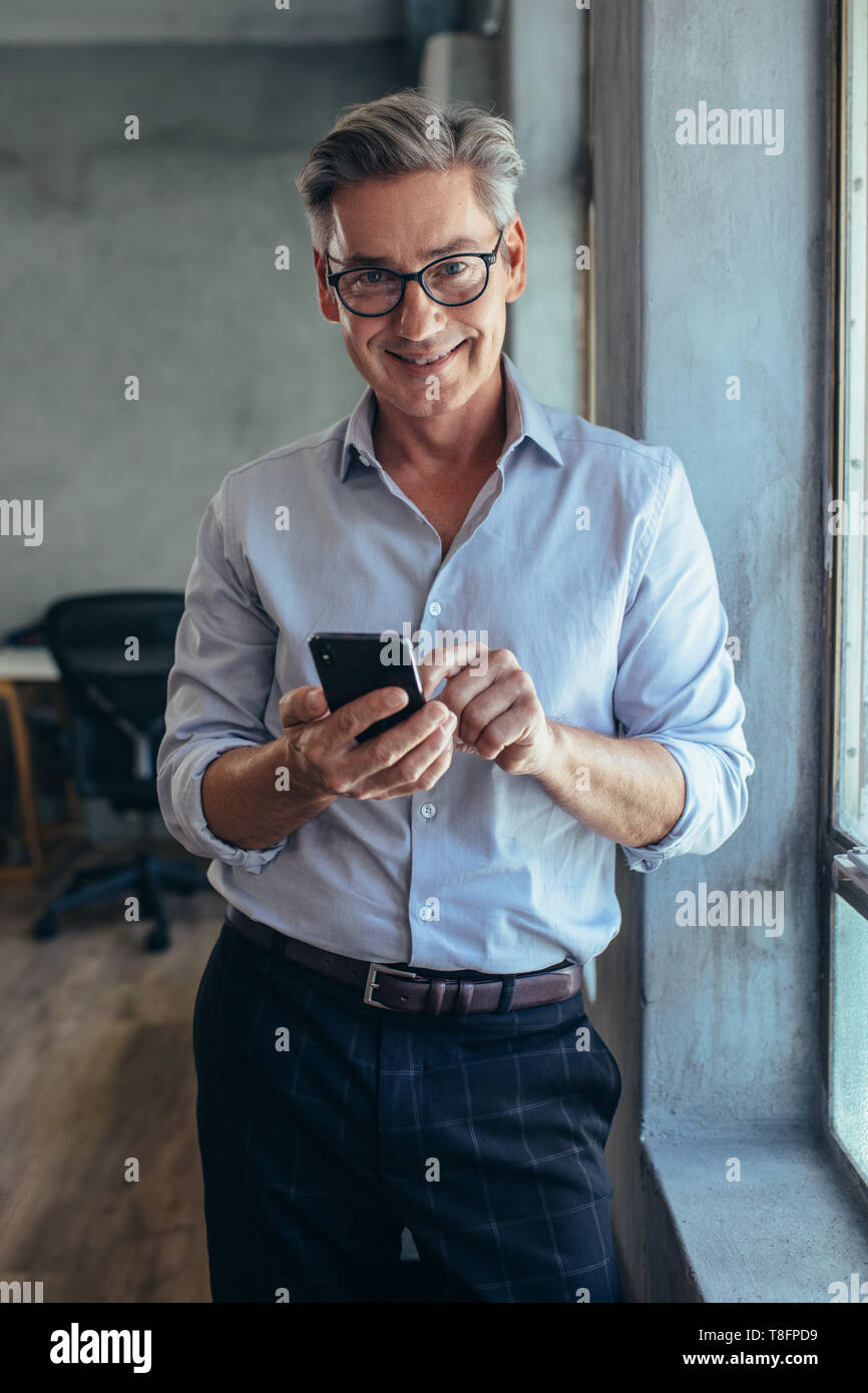 Happy businessman matura in piedi da una finestra con il suo telefono cellulare. Imprenditore maschio nel suo ufficio. Foto Stock