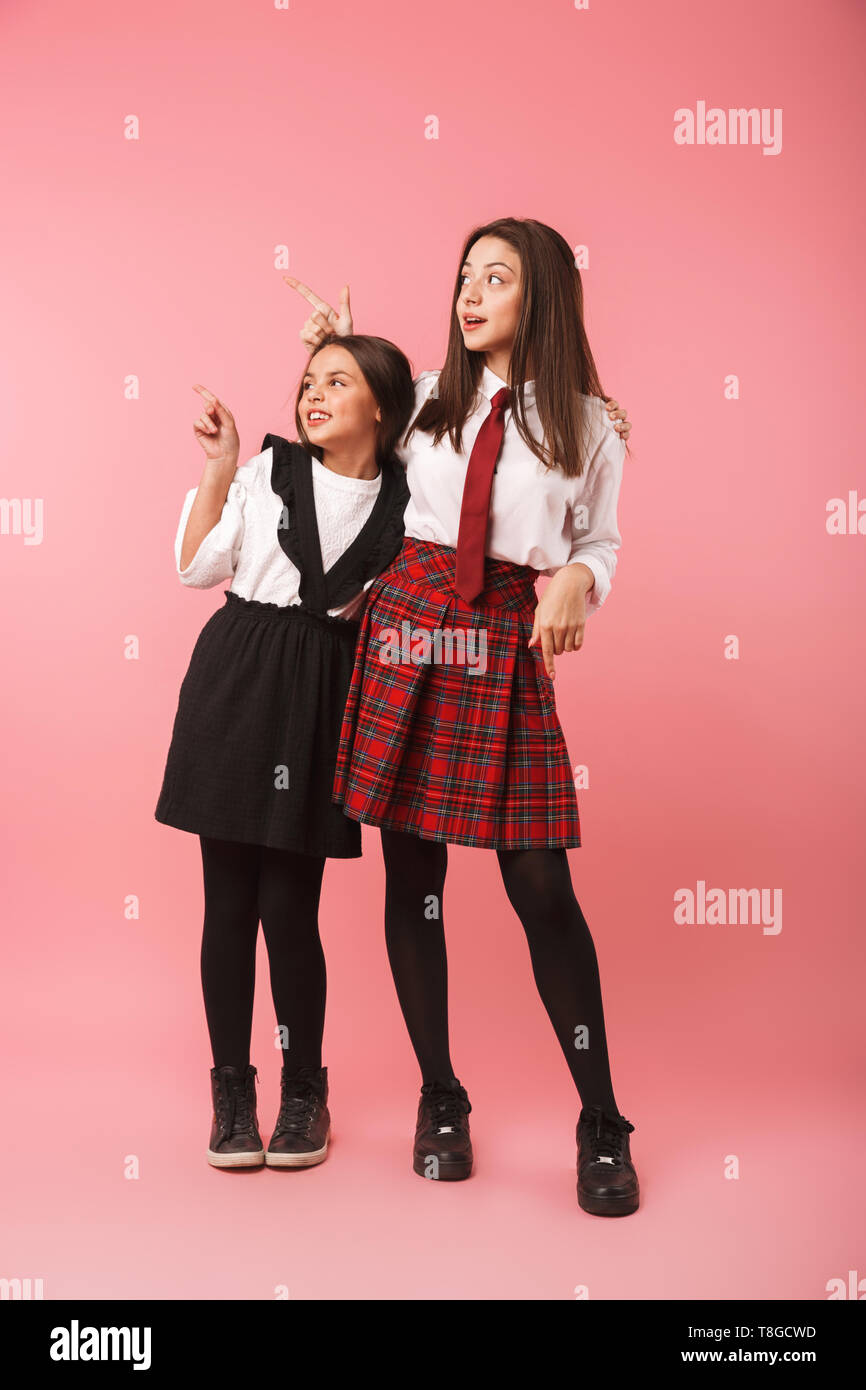 Due allegro scuola ragazze indossano uniformi isolato permanente su sfondo rosa, dita di puntamento Foto Stock