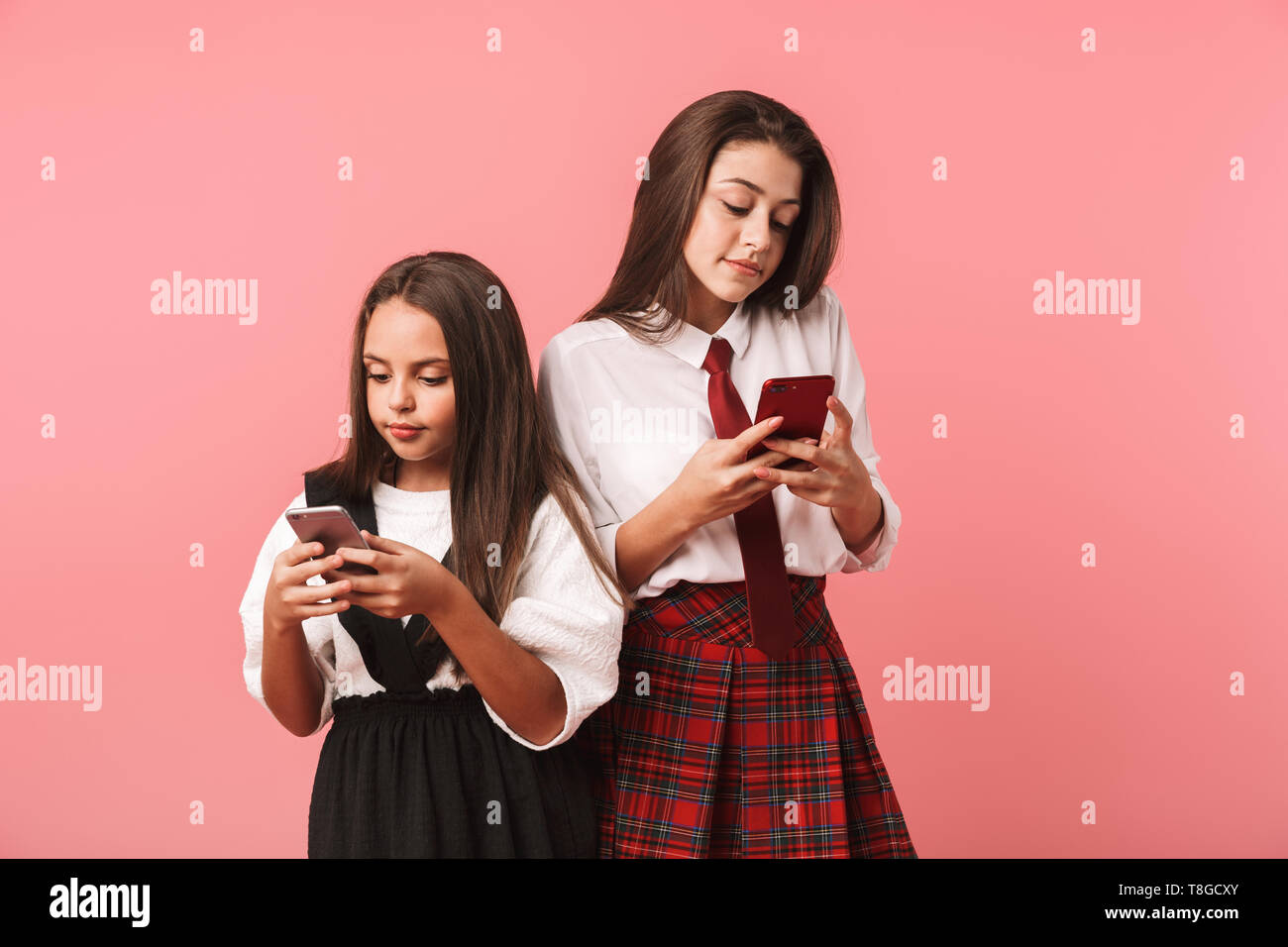 Ritratto di belle ragazze in uniforme scolastica utilizzando gli smartphone in piedi mentre isolate su sfondo rosso Foto Stock