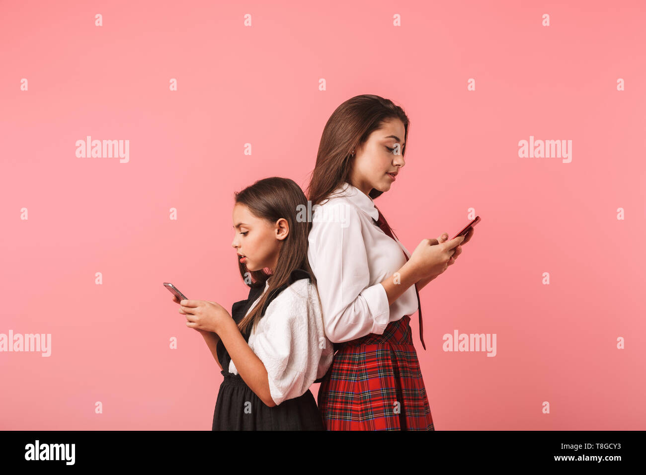 Ritratto di bruna ragazze in uniforme scolastica utilizzando gli smartphone in piedi mentre isolate su sfondo rosso Foto Stock