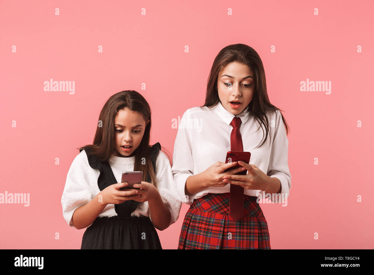 Ritratto di ragazze eccitato in uniforme scolastica utilizzando gli smartphone in piedi mentre isolate su sfondo rosso Foto Stock