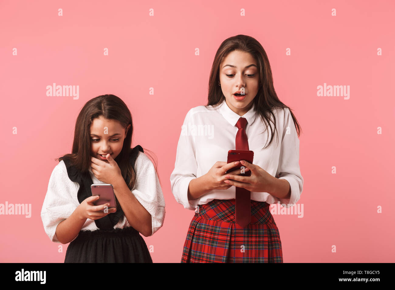 Ritratto di ottimista ragazze in uniforme scolastica utilizzando i telefoni cellulari mentre in piedi isolato su sfondo rosso Foto Stock