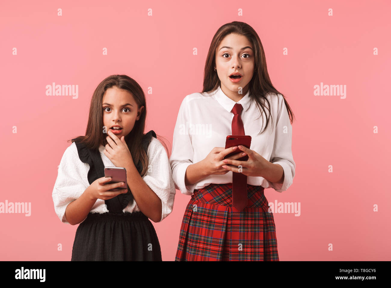 Ritratto di ragazze positivo in uniforme scolastica utilizzando i telefoni cellulari mentre in piedi isolato su sfondo rosso Foto Stock