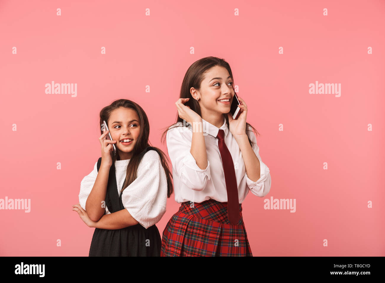 Ritratto di belle ragazze in uniforme scolastica utilizzando i telefoni cellulari per le chiamate mentre in piedi isolato su sfondo rosso Foto Stock