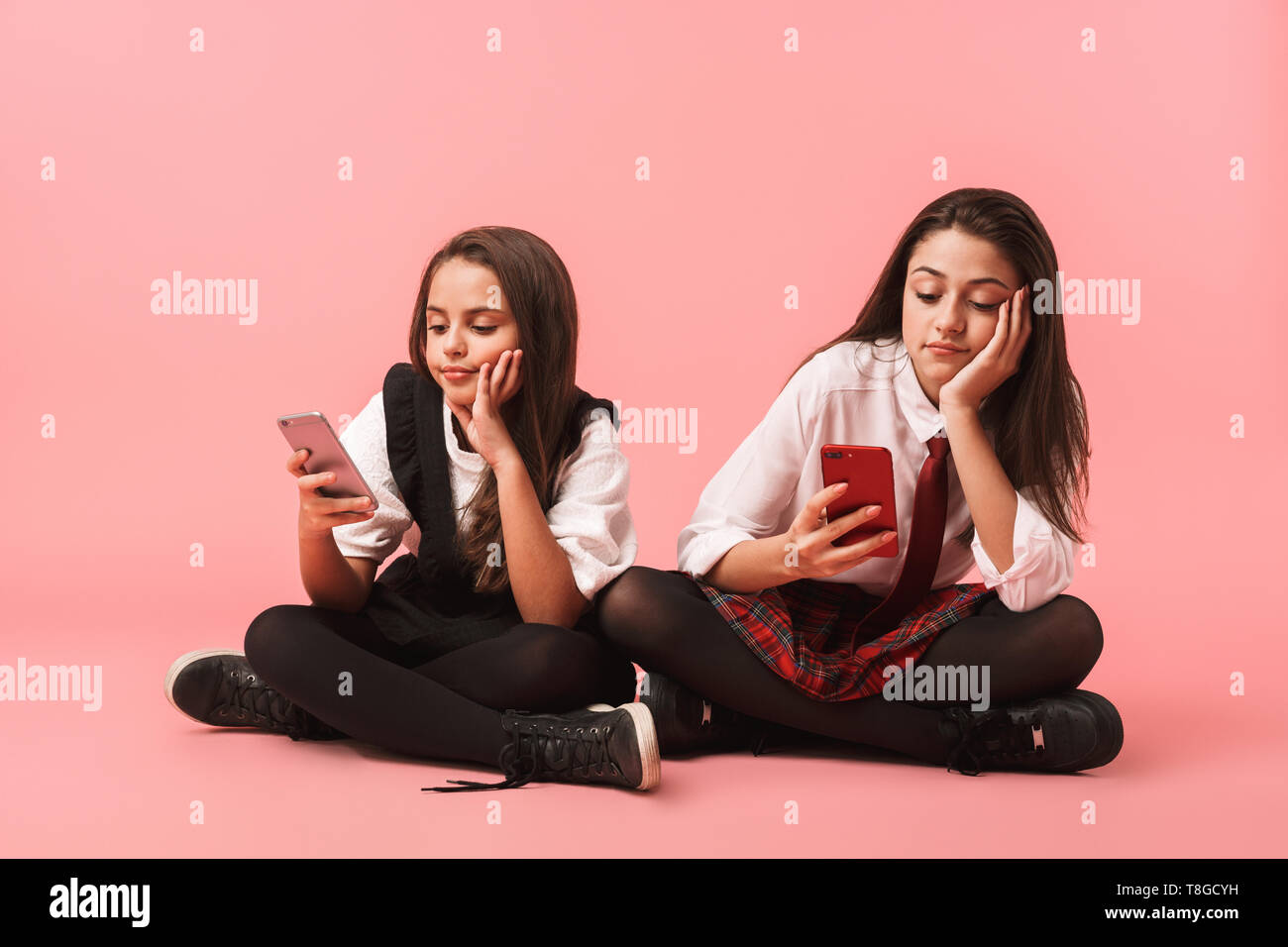 Ritratto di belle ragazze in uniforme scolastica utilizzando i telefoni cellulari mentre è seduto sul pavimento isolato su sfondo rosso Foto Stock