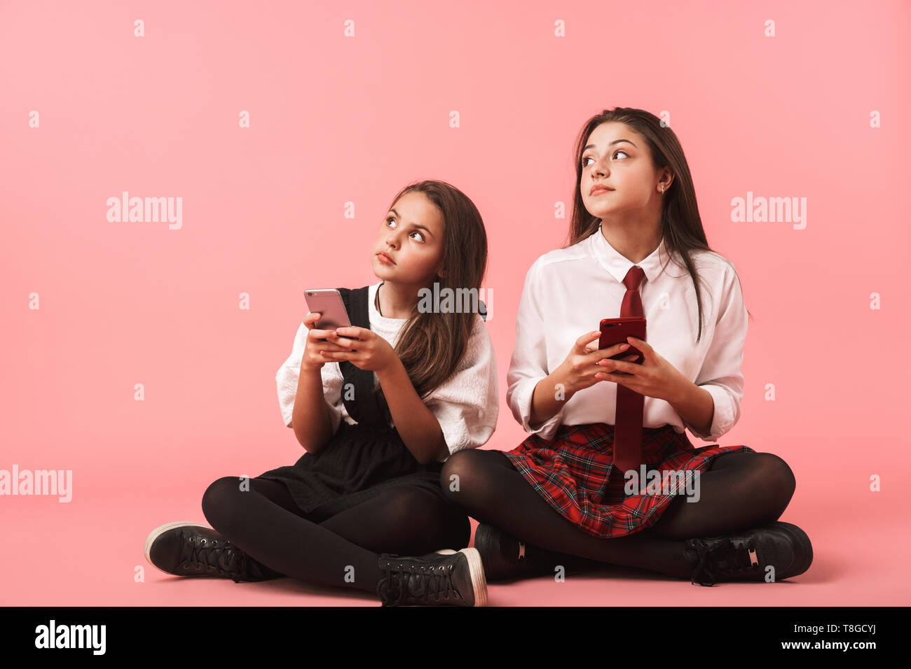 Ritratto di Allegro ragazze in uniforme scolastica utilizzando i telefoni cellulari mentre è seduto sul pavimento isolato su sfondo rosso Foto Stock