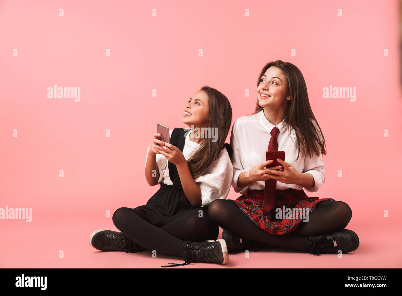 Ritratto di giovani ragazze in uniforme scolastica utilizzando i telefoni cellulari mentre è seduto sul pavimento isolato su sfondo rosso Foto Stock