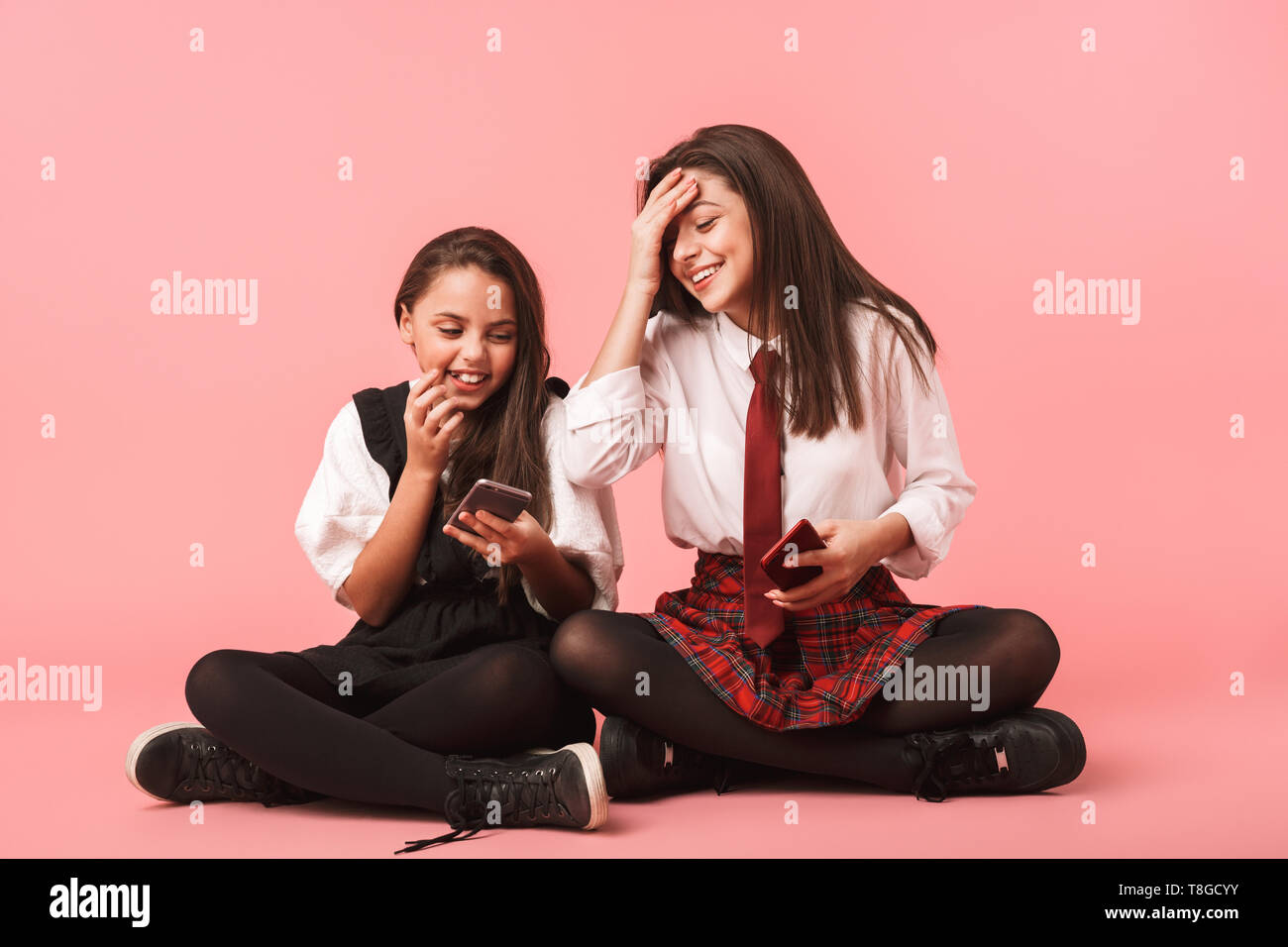 Ritratto di ragazze sorridenti in uniforme scolastica utilizzando i telefoni cellulari mentre è seduto sul pavimento isolato su sfondo rosso Foto Stock