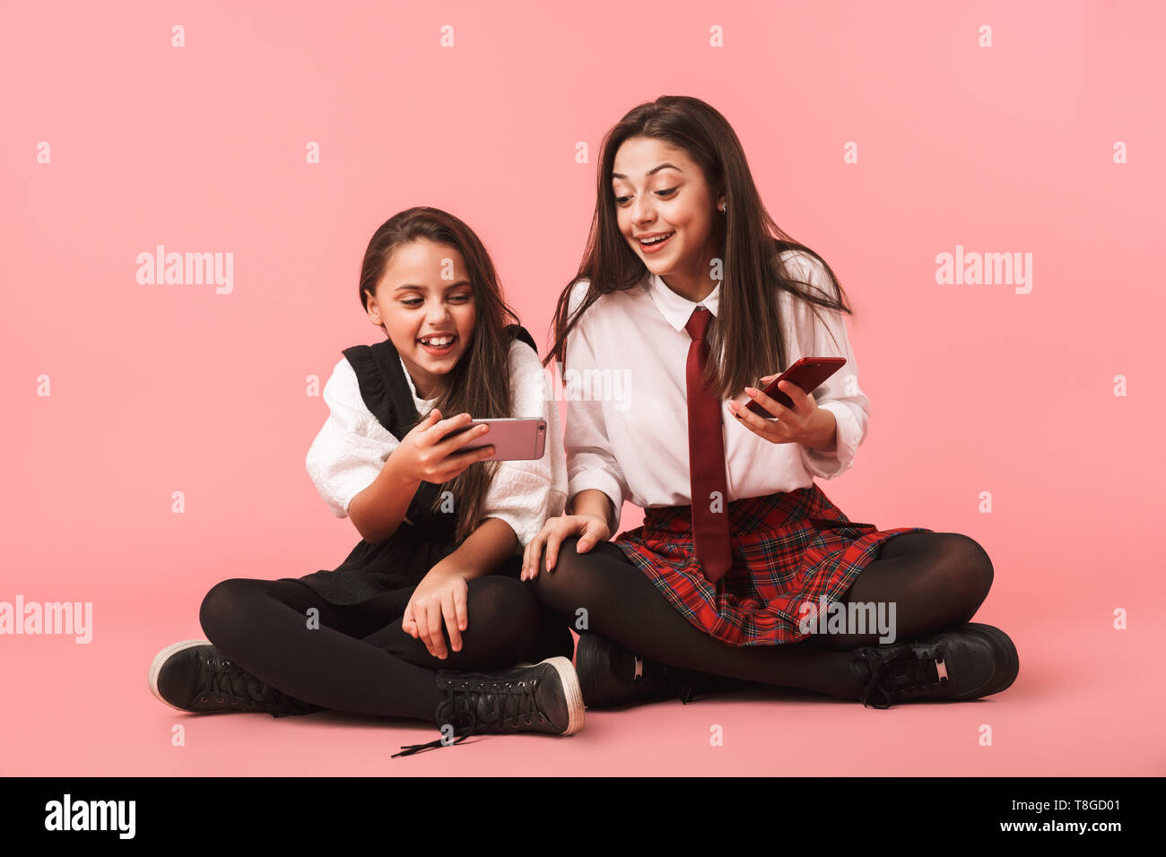 Ritratto di gioiosa ragazze in uniforme scolastica utilizzando i telefoni cellulari mentre è seduto sul pavimento isolato su sfondo rosso Foto Stock