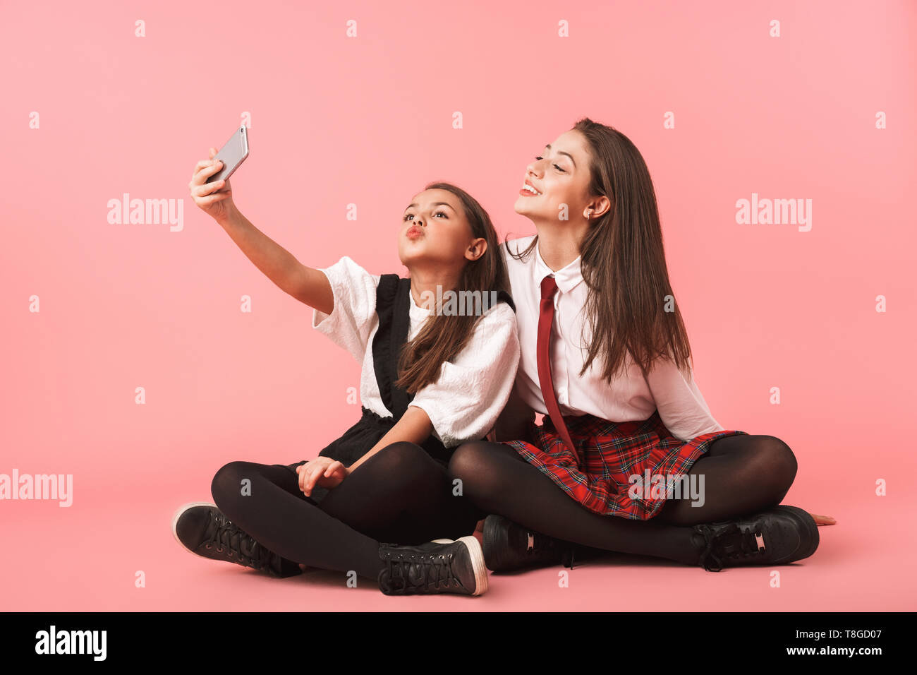 Ritratto di divertenti ragazze in uniforme scolastica utilizzando i telefoni cellulari per selfie foto mentre è seduto sul pavimento isolato su sfondo rosso Foto Stock