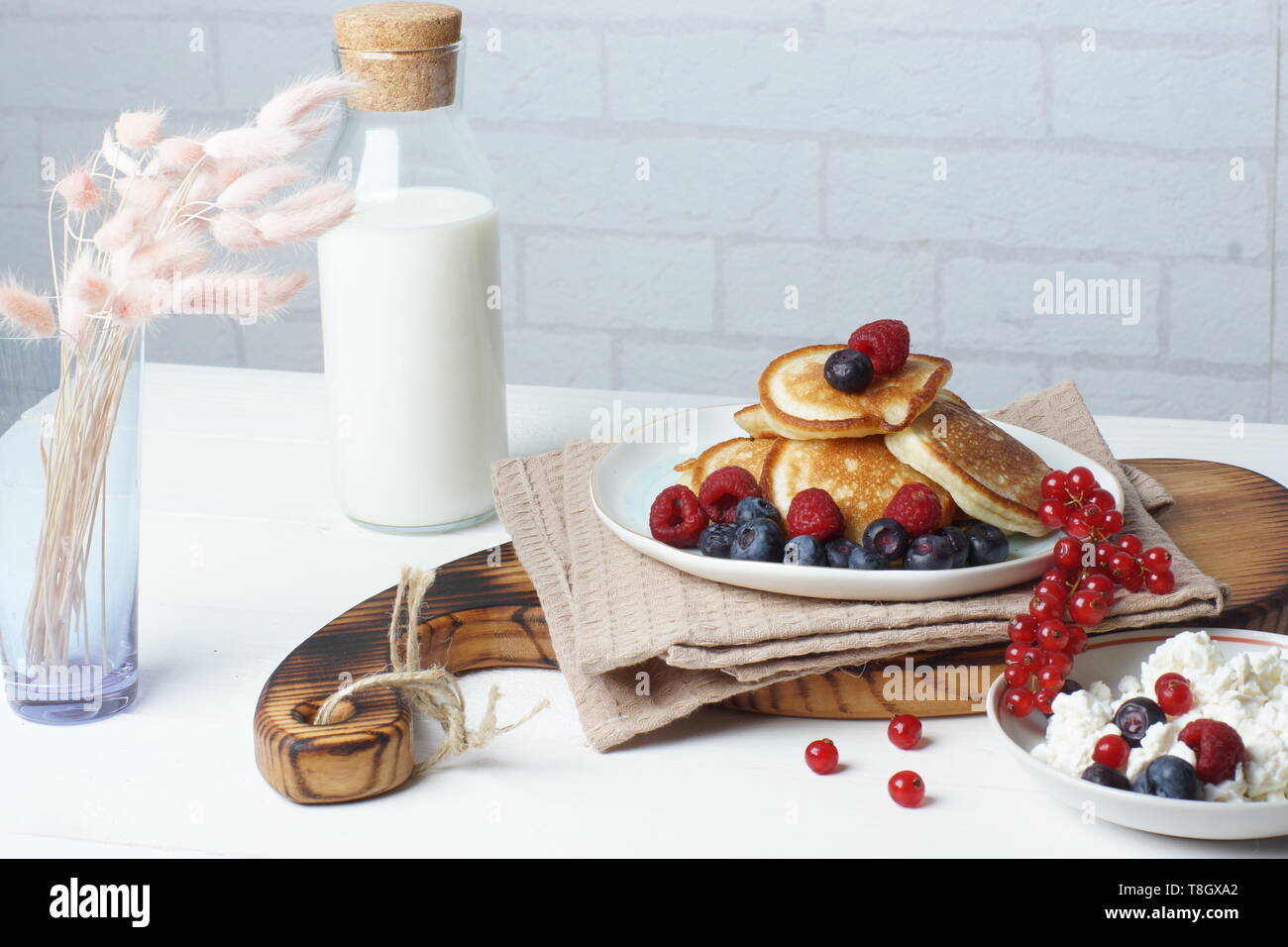 La colazione su un tavolo bianco, frittelle con frutti di bosco freschi, formaggio e una bottiglia di latte Foto Stock