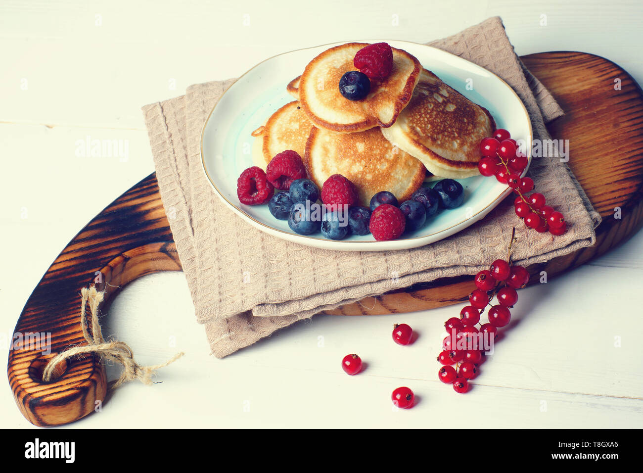 Su un tavolo bianco si erge una tavola di legno con colazione, appena sfornato pancake con frutti di bosco freschi di ribes, mirtilli e lamponi. Foto Stock