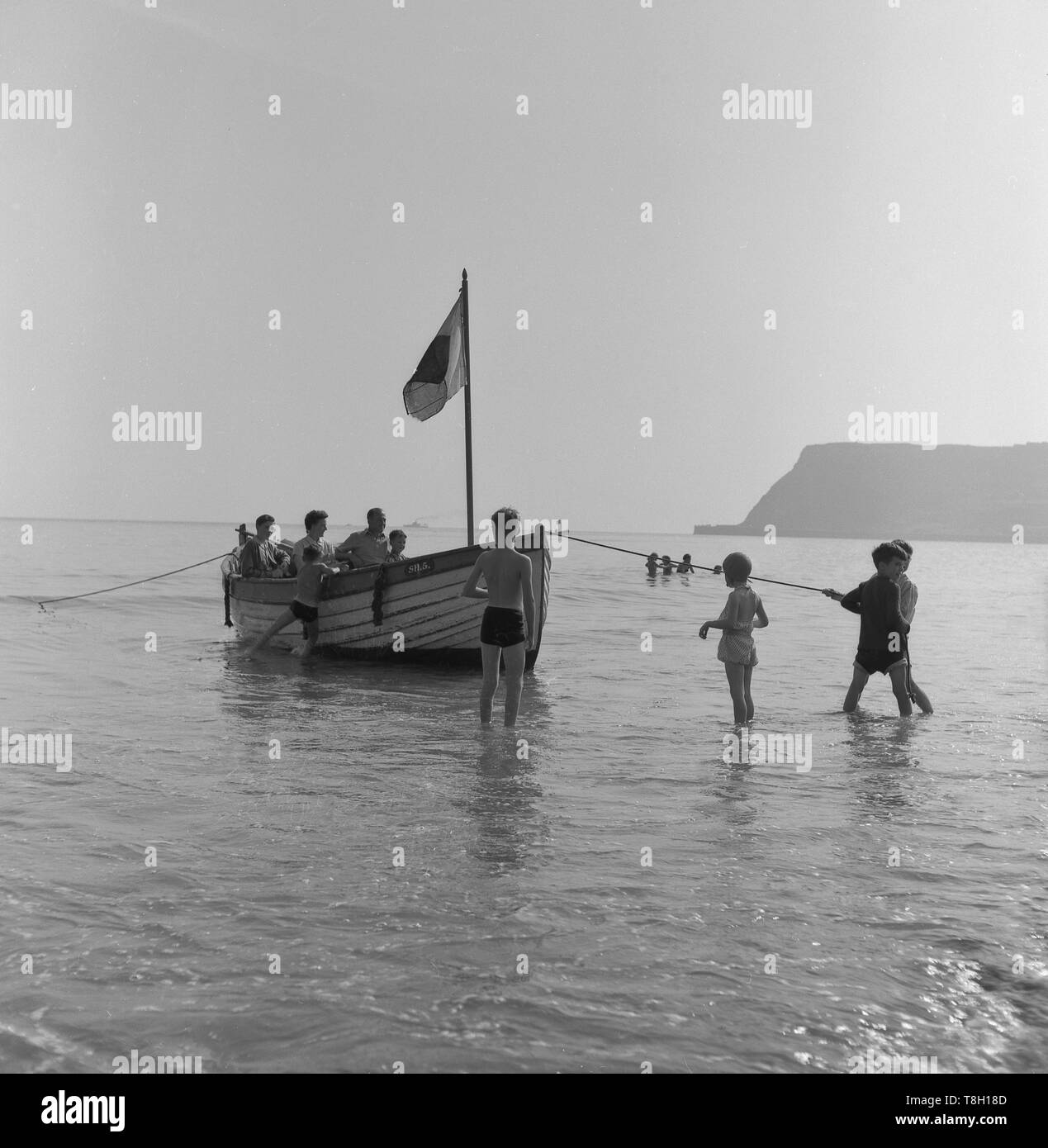 Degli anni Cinquanta, storico in legno barca a remi in acqua poco profonda da mari edge, ragazzi giovani in possesso di una fune per tirare la barca in riva, Inghilterra, Regno Unito. Foto Stock